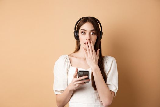 Surprised young woman gasping and covering mouth with hand, using wireless headphones to listen podcast or music, holding mobile phone, beige background.