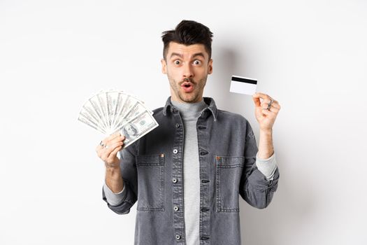 Excited guy holding plastic credit card and dollar bills, standing amused on white background.