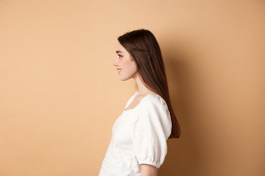 Profile of beautiful caucasian woman with long natural hair, looking left at empty space and smiling romantic, standing on beige background.
