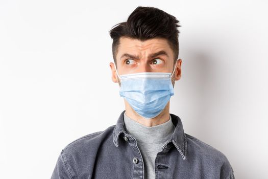 Pandemic lifestyle, healthcare and medicine concept. Confused guy in face mask look left at logo and frowning skeptical, standing on white background.