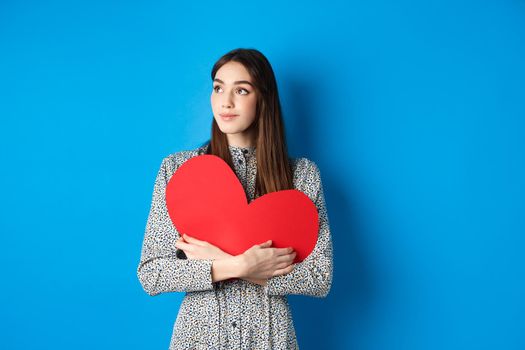 Valentines day. Romantic girl looking dreamy at upper left corner and smiling, holding big red heart cutout, standing on blue background.