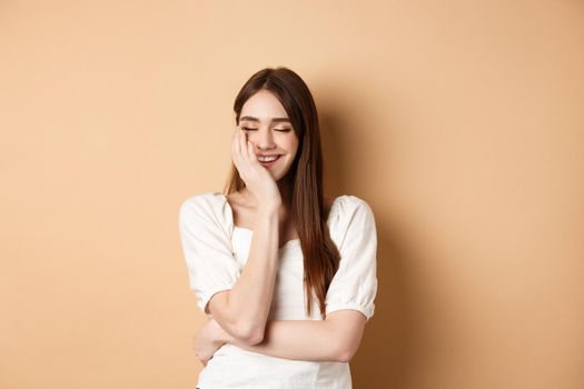 Happy and romantic girl laughing with eyes closed, touching face and feeling joy, standing on beige background.