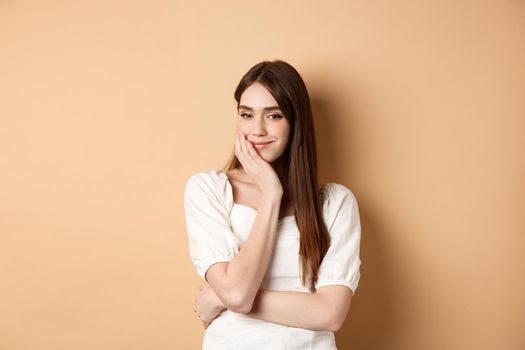 Romantic young woman touching face and smiling at camera with pensive look, standing dreamy on beige background.