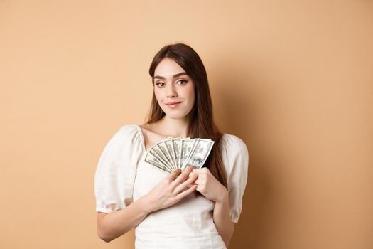 Smiling rich woman showing dollar bills and looking happy at camera, making money or fast loan cash, standing on beige background.