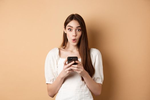 Girl standing amazed with smartphone, saying wow and look excited by awesome online offer news, standing on beige background.