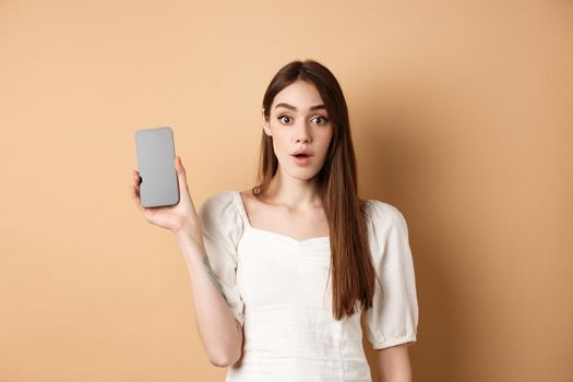 Excited pretty girl showing empty cell phone screen, look amazed at camera, standing on beige background.