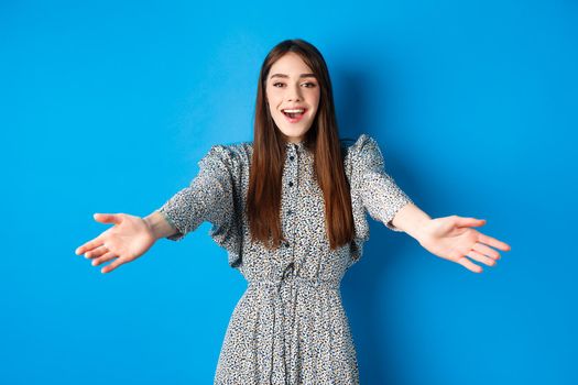 Beautiful happy woman in dress stretch out hands in warm welcome, smiling and greeting you, inviting guests, standing on blue background.