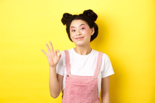 Cheerful asian beauty girl with makeup, show okay sign and smiling excited, approve good promo, recommending product, yellow background.