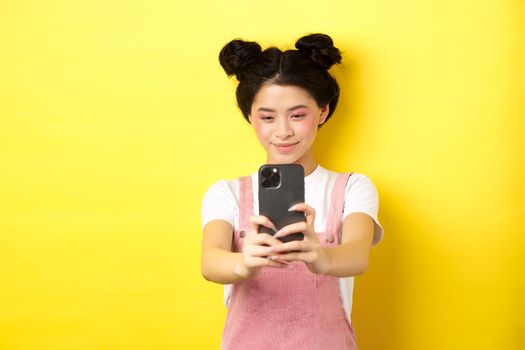 Stylish asian girl taking photo on smartphone, making video with cellphone and smiling, standing on yellow background.