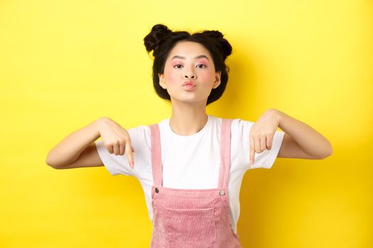 Sassy asian teen girl pucker lips and pointing fingers down, tell to look at bottom advertisement, standing with glamour makeup and summer clothes on yellow background.