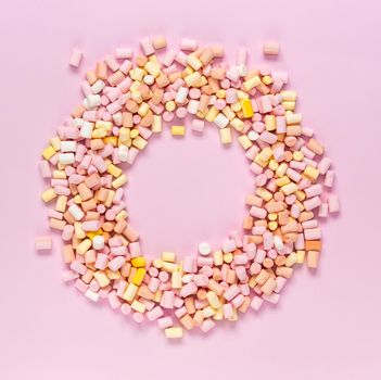 Top view of the multi-colored marshmallows which lies in the shape of a round frame with an area for text in the center on a monochrome pink background.