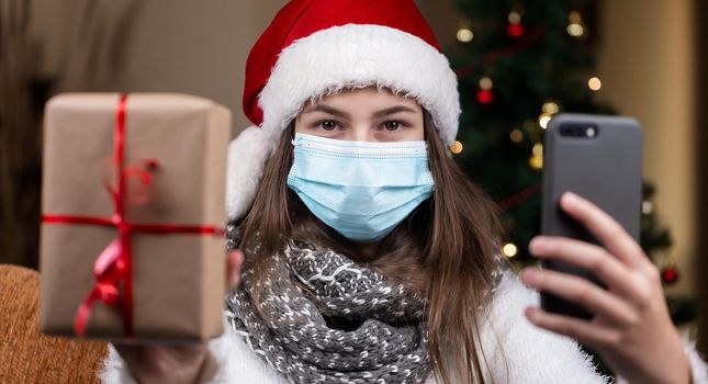 Christmas online greetings. A young girl in a santa claus hat and a medical mask talks using smartphone for video call friends and parents. Christmas during the coronavirus