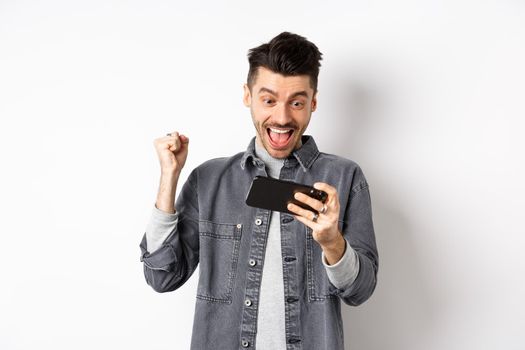 Happy man shouting yes, winning video game on mobile phone and triumphing, achieve online goal, standing on white background.