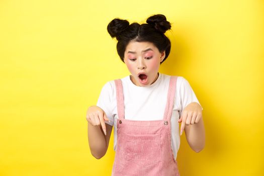 Excited teenage asian girl checking out advertisement, pointing fingers down and drop jaw amazed, showing promo offer, yellow background.