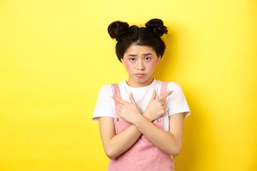 Indecisive timid asian girl pointing sideways, showing left and right ways and sulking upset, standing sad on yellow background.