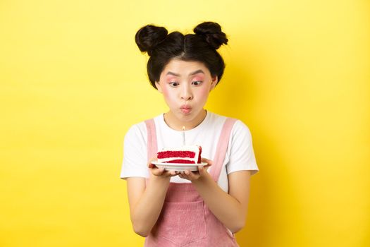 Holidays and celebration. Silly asian girl with glamour makeup, making wish and blowing candle on birthday cake, standing on yellow background.