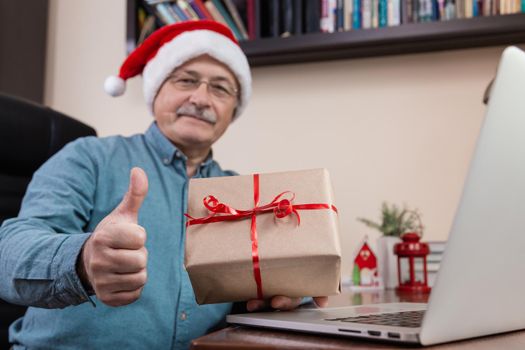 Christmas online congratulation. Senior man in santa claus hat Gives a gift and talks using laptop device for video call friends and childrens. Christmas during coronavirus.