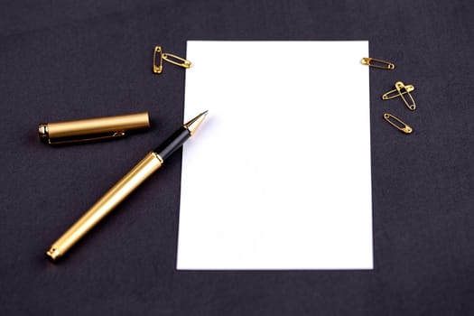 Gold pen, ribbon, paper clips and stationery on a black background with a white sheet of paper.