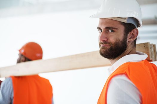 Two young men builders carrying wood planks on construction site, close up photo