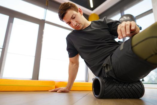 The man is massaging his leg muscles with the foam roller in gym