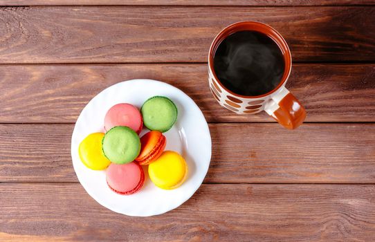 Colorful sweet macarons dessert on a plate and cup of hot drink on wooden table, top view.