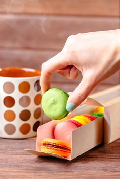 Female hand holding macarons near the cup of hot drink on wooden background.