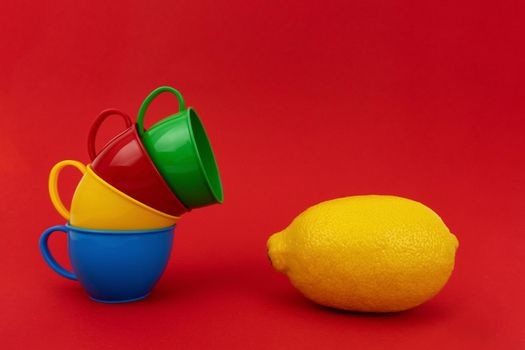 Four colourful small mugs, green, red, yellow, blue, stay one in another near yellow lemon on red background. Enough space for text. Minimalistic concept.