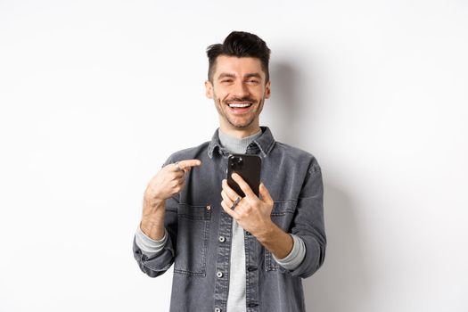 Happy handsome caucasian man with moustache pointing finger at smartphone, smiling and showing good deal online on mobile phone, standing pleased against white background.