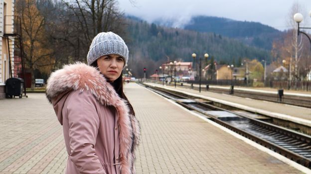 A woman in a gray knitted hat and a pink jacket with fur is waiting for the train at the station looking at the arriving train. Rail transportation. Autumn and winter travel concept