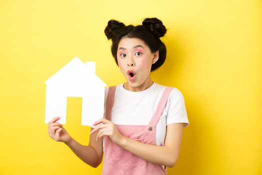 Real estate and family concept. Excited asian teen girl showing paper house cutout and gasping amazed, standing on yellow background.