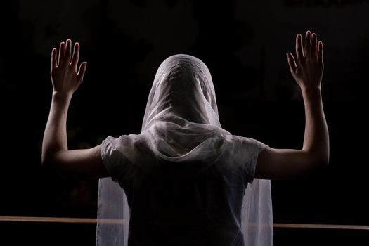 Silhouette of a young girl who sits in church and praying with hands raised up. Close-up view from behind. Backlight.