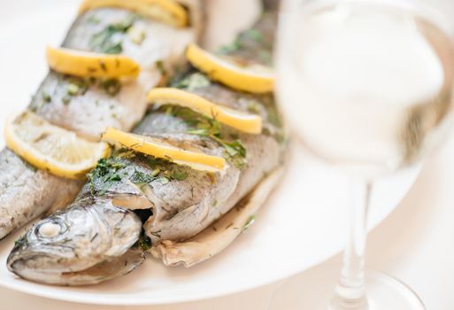 Fish dish with lemons and greenery on a white plate with glass of white wine for dinner.
