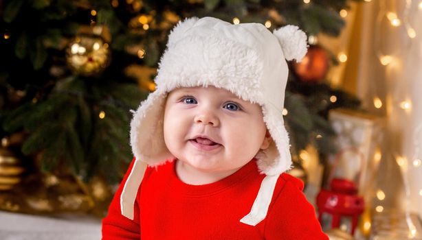 Christmas baby is smiling. A cute little girl in a red dress and white hat expresses emotions. Christmas concept with little kid, tree and garland on background in blur