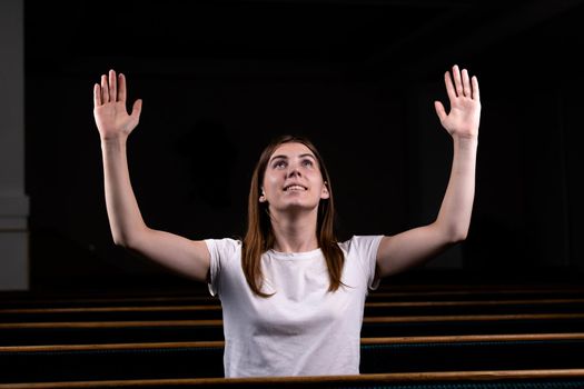 A Christian girl in white shirt is sits with his hands up and face and praying with humble heart in the church.