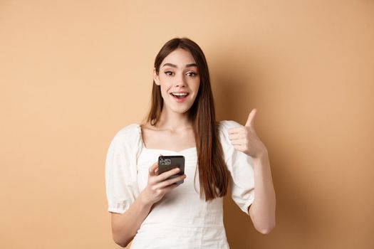 Online shopping concept. Happy young woman showing thumb up and using cellphone, smiling pleased, standing on beige background.