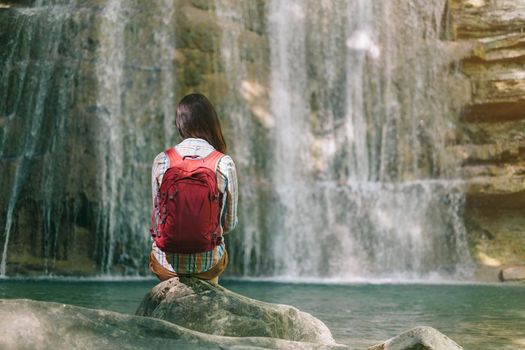 Traveler explorer young woman with backpack sitting on stone and enjoying view of waterfall in summer.