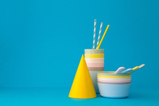 Composition with multicolored plastic reusable tableware with straws and bright yellow party hat on blue background