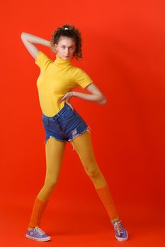 Pretty fitness girl posing relaxed on red backdrop. Girl with ponytail girl standing with legs wide apart, one hand behind her head and other at waist