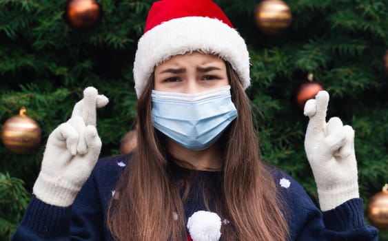 holding fingers crossed for good luck at Christmas. Close up Portrait of woman wearing a santa claus hat and medical mask with emotion. Against the background of a Christmas tree. Coronavirus pandemic