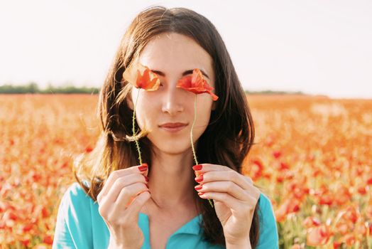 Beautiful brunette young woman holding red poppies in front of her eyes and standing in flower meadow.