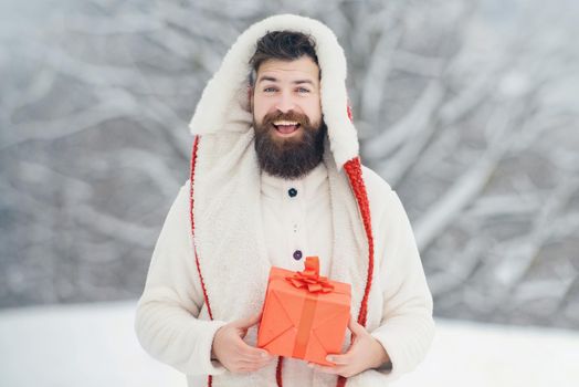 Christmas winter portrait of handsome hipster in snow with gift make snowman. Man with red gift. Bearded handsome man, Santa Claus with beard.