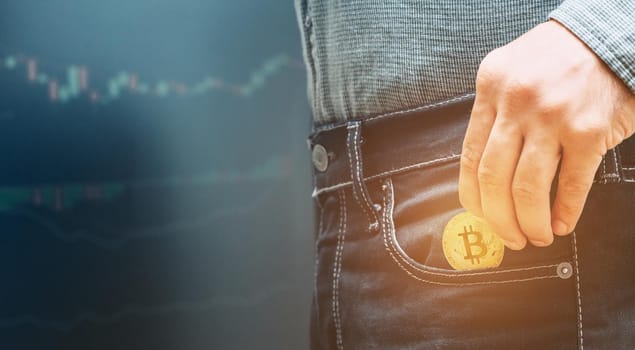 Unrecognizable man holding gold bitcoin and putting into pocket of jeans on background of infographics.