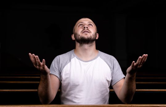 A Christian man in white shirt is sitting with his hands up and praying with humble heart in the church.