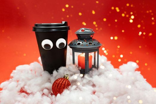 Black coffee cup with plastic cap and googly eyes in snow near night lantern on red background with lights, bokeh style, new year concept, place for the text.