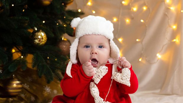 Christmas child looking at the camera and holding a garland with hearts. A cute little girl in a red dress and white hat. Christmas concept with little kid, tree and garland on background in blur