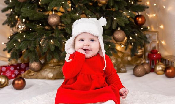 Christmas baby looking at the camera. A cute little girl in a red dress and white hat expresses emotions. Christmas concept with little kid, tree and garland on background in blur