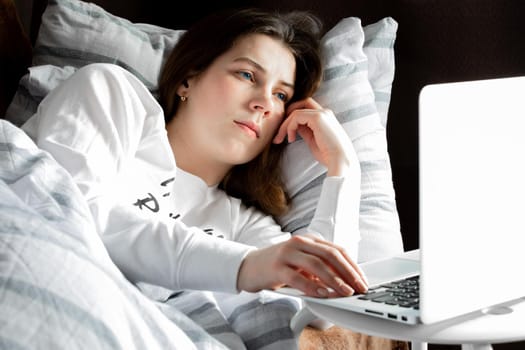 Young cute girl on a sunny day in a white jacket lies on the bed and looks into the laptop.