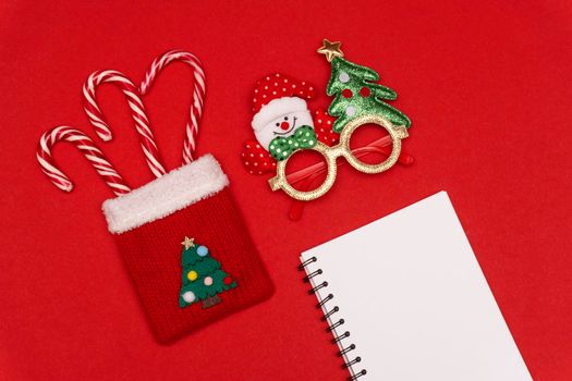 Flatlay at Christmas bag with striped candies, Christmas glasses and white sheet of notebook for future 2021 plans and motivation, red background, copyspace, Christmas and new year concept.