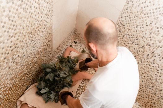 Slim woman in bath is getting steaming massage with hot oak leaves brooms, in steam-room.