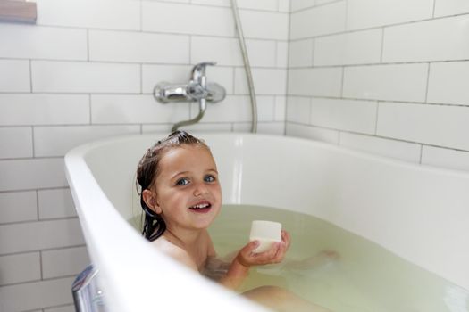 Positive little girl sitting in water of bath with bar soap while washing in bathtub and looking away